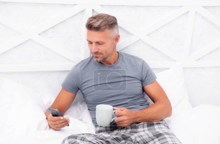 Photo for Smiling man messaging in morning. man messaging in morning in bed. man messaging in morning in bedroom. photo of man messaging in morning at home. - Royalty Free Image