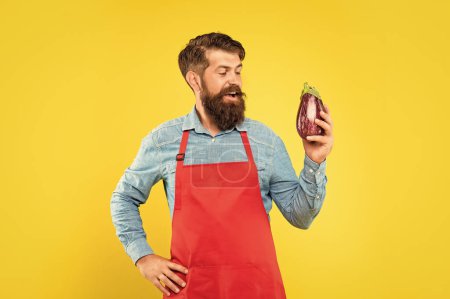 Happy man in red apron looking at eggplant yellow background, grocer.