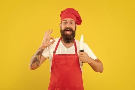Glücklicher Mann in roter Schürze und Haube gibt OK halten Köche Messer gelben Hintergrund, Koch.