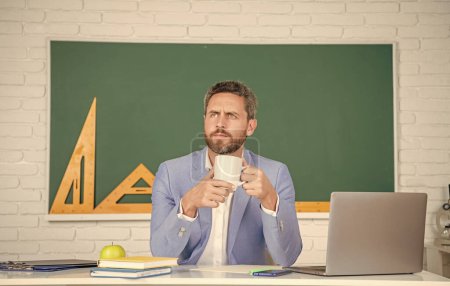 thoughtful school teacher in classroom with laptop at blackboard.