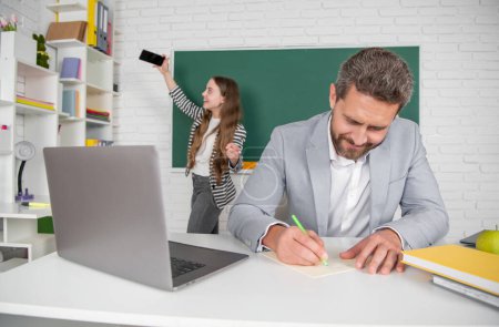 smiling school teacher in classroom with selective focus of kid making selfie at blackboard.