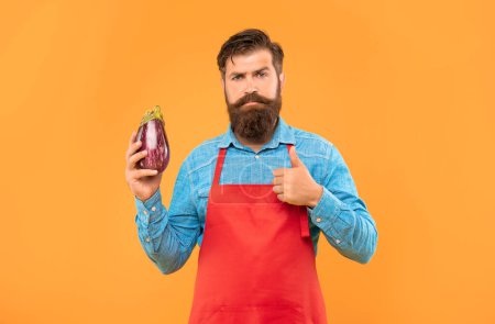 Serious man in red apron giving thumb gesture to eggplant yellow background, greengrocer.