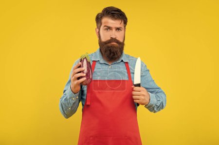 Ernsthafter Typ in Schürze mit Kochmesser und Auberginen-gelbem Hintergrund, Koch.