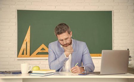 school teacher in classroom with laptop at blackboard.