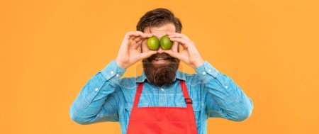 Hombre feliz en delantal sosteniendo limas frescas sobre los ojos fondo amarillo, vendedor de frutas.