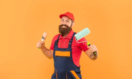happy bearded man painter in work clothes hold paint roller and brush on yellow background.