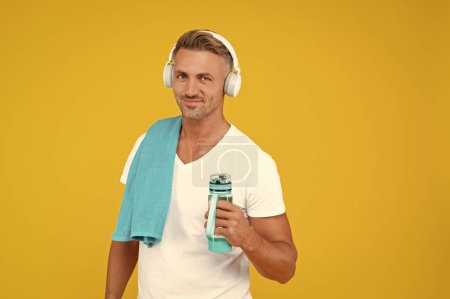 happy guy hold bottle for healthy lifestyle. guy with sport bottle for healthy lifestyle isolated on yellow. guy holding bottle to keep sport lifestyle. guy with towel and bottle after training.