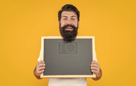 studio advertisement of smiling man. bearded man at advertisement blackboard with copy space. bearded man with advertisement blackboard isolated on yellow. man hold blackboard for advertisement.