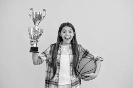 surprised teen basketball girl receive award in studio. teen basketball girl accept award. proud teen basketball girl with champion cup award. teen basketball girl hold award cup isolated on yellow.