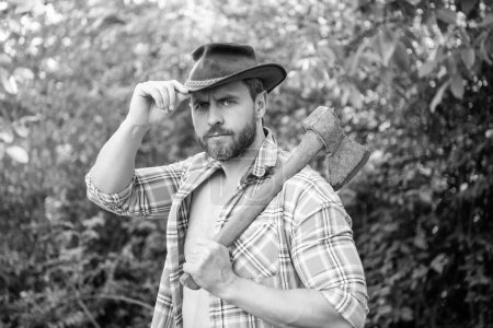 cowboy with axe in hat outdoor. photo of cowboy with axe. cowboy with axe. cowboy with axe wearing checkered shirt.