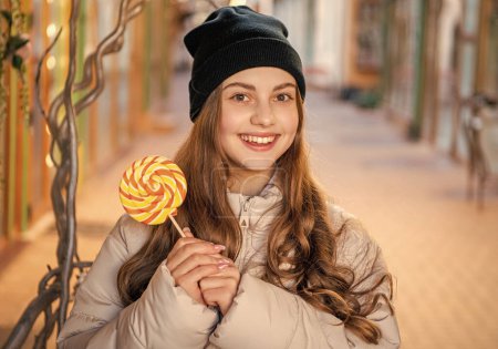 fröhliches Mädchen mit Lollypop-Bonbons auf der Straße. Mädchen mit Lollypop-Süßigkeiten im Freien. Mädchen mit Lollypop-Bonbons mit Hut. Foto von Mädchen mit Lollypop Süßigkeiten draußen.