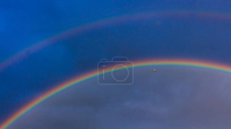 Foto de El avión vuela bajo el arco iris. Un arco iris en el cielo azul. Siete colores del arco iris. - Imagen libre de derechos