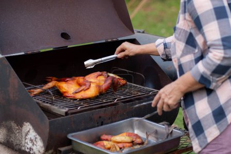 Foto de Senior woman grilling chicken meat for preparing eat in camping, elderly doing activity recreation and leisure picnic while roasting meat in weekend, travel trip in holiday, party in vacations. - Imagen libre de derechos