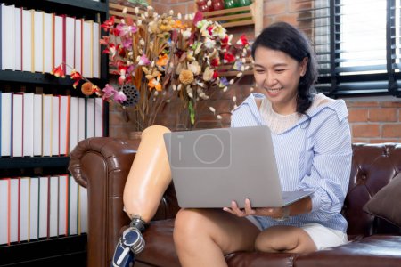 Photo for Young asian woman disabled sitting on sofa using laptop computer with prosthetic leg in living room at home, female working on notebook and prosthesis with optimistic, lifestyles and disability. - Royalty Free Image