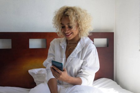 Foto de Chica hispana joven usando el teléfono celular en la cama sonriendo - Imagen libre de derechos