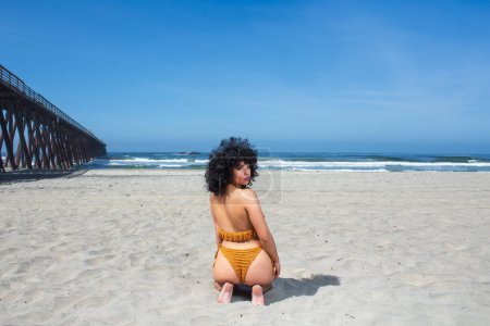 Foto de Hermosa y sensual joven afroamericana mexicana sentada en la playa de rosarito al lado del muelle mirando a la cámara, vistiendo bikini amarillo en verano - Imagen libre de derechos