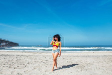 Foto de Hermosa mujer afroamericana en la playa de Rosarito mexicana con pantalones cortos y bikini de pie junto al muelle, latina - Imagen libre de derechos