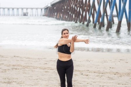 Foto de Deportista, mujer latina de mediana edad calentando y estirando los brazos en la playa, madura mexicana fitness - Imagen libre de derechos