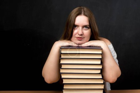Foto de Una mujer de apariencia eslava se sienta en una mesa con la cabeza apoyada en una pila de libros. fondo negro. un profesor o un estudiante se está preparando para un examen - Imagen libre de derechos