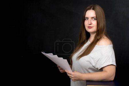 Foto de Una chica de apariencia europea sostiene papeles en sus manos sobre un fondo negro. profesor u oficinista - Imagen libre de derechos