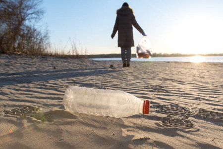 Foto de Botella de plástico en la orilla del río. cuestiones ambientales y el día de la tierra. chica voluntaria en el fondo con una bolsa de basura - Imagen libre de derechos