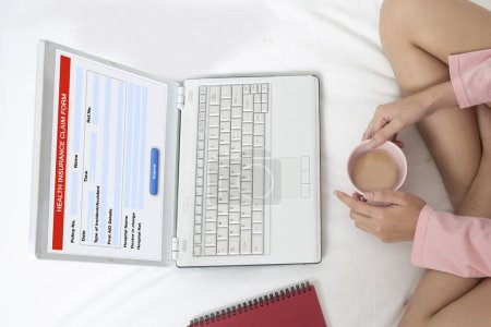 Photo for High angle photo of young woman sits on the bed and fills out health insurance claim form that display on notebook computer. - Royalty Free Image