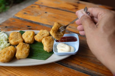 Photo for Someone hand uses fork to get a picec of chicken nuggets from disk on wooden table. - Royalty Free Image