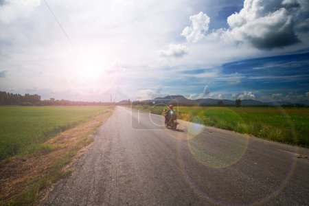 Téléchargez les photos : Une personne roule à moto devant sur une route de campagne dans un pays asiatique en passant par un vaste champ de riz sous le soleil ardent seul. - en image libre de droit