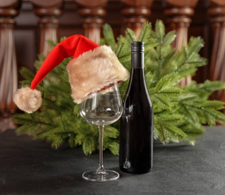 Téléchargez les photos : Une bouteille de vin, un verre avec un chapeau de Père Noël sur un fond de branches de sapin. Composition de Noël. - en image libre de droit