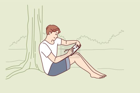 Man draws in electronic tablet sitting on shore of lake and enjoying beautiful landscape of morning nature. Creative young man is experiencing surge of inspiration being in park with river