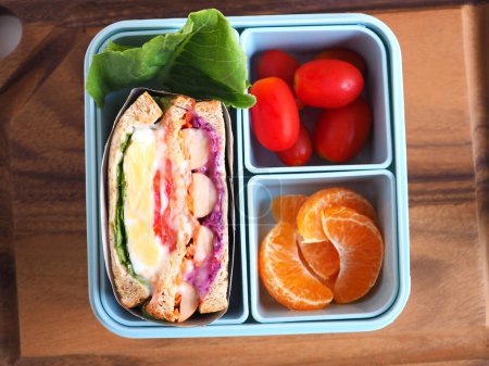 Homemade healthy blue lunch box with sandwich and orange, tomato vegetables, on wooden background. Top view, with copy space.
