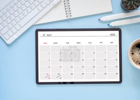 The online calendar app on the tablet computer showing screen July 2023, on blue background and keyboard, notebook, cactus. Reminder with the calendar application for schedule planning. Top view