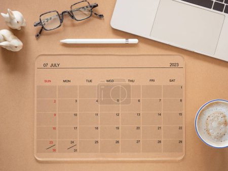 Draufsicht, flache Lage, der Online-Kalender auf einem transparenten Tablet-Computer mit Bildschirm Juli 2023, auf braunem Hintergrund, Kaffeetasse, Laptop und Brille. Mahnung zur Terminplanung