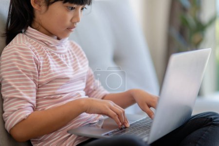 Téléchargez les photos : Asiatique petite écolière utiliser ordinateur portable assis sur canapé seul à la maison. Apprendre à lire des contenus de médias sociaux en ligne, jouer à des leçons d'éducation jeu bavarder avec des amis. Dépendance technologique des enfants - en image libre de droit