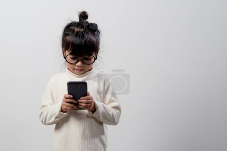 Photo for Asian little girl  using a phone close up, distracted from studying, sitting at a table with notebooks, a pretty child having fun with a smartphone, watching the webinar, homeschooling - Royalty Free Image