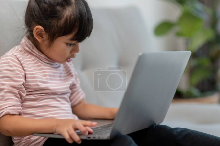 Téléchargez les photos : Asiatique petite écolière utiliser ordinateur portable assis sur canapé seul à la maison. Apprendre à lire des contenus de médias sociaux en ligne, jouer à des leçons d'éducation jeu bavarder avec des amis. Dépendance technologique des enfants - en image libre de droit