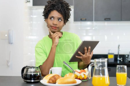 Téléchargez les photos : Thinking and dreaming african american female student with tablet computer indoors at home at breakfast - en image libre de droit