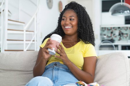 Téléchargez les photos : Rire corpulent afro-américaine jeune adulte buvant du café à l'intérieur à la maison - en image libre de droit