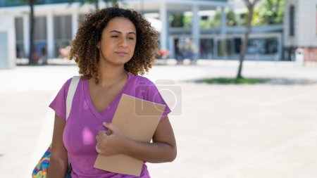 Estudiante hispana seria con mochila y papeleo frente a la universidad al aire libre en verano en la ciudad