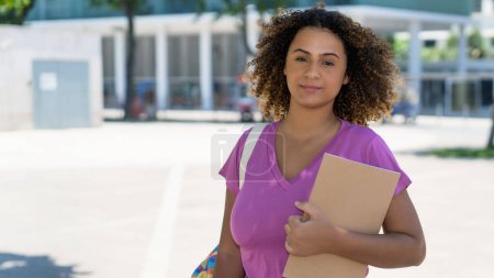 Hispanische Studentin mit Rucksack und Papierkram vor der Universität im Sommer in der Stadt