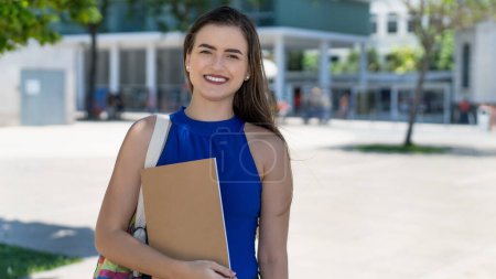 Riendo joven caucásica estudiante con pelo morena al aire libre delante de la universidad en verano