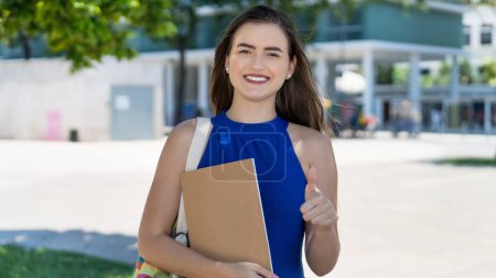Successful young caucasian female student with brunette hair showing thumb up outdoor in front of university in summer