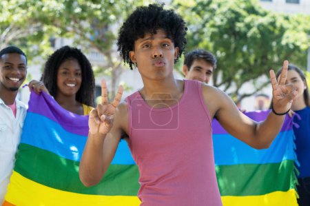 Funny black male young adult celebrating pride parade with LGBTQ rainbow flag and group of queer people outdoor in summer in city