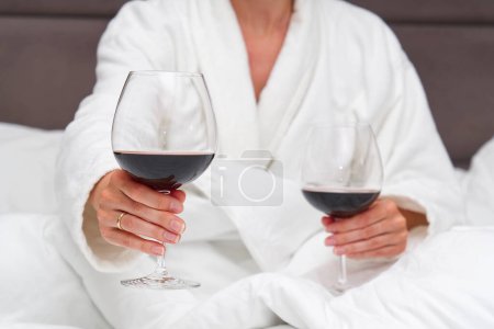 Photo for Unrecognizable cute pair of hands holding flasks with red wine in bed. romance. Lying in bed in cozy hotel - Royalty Free Image