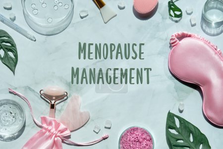 Menopause management text on wellness flat lay. Pink stone facial roller and guasha stone on mint green with monstera leaves. Towel, sleep mask, moisturizer in glass petri dish.