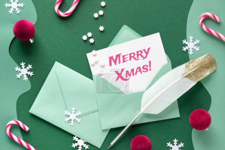 Photo for Writing Christmas greetings - quill and greeting cards in paper envelopes. Xmas background with candy canes, trinkets, snowflakes. Flat lay on green paper, greeting text caption Merry Xmas. - Royalty Free Image
