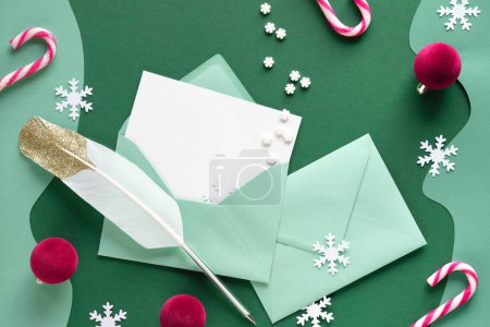 Photo for Writing Christmas greetings - quill and greeting cards in paper envelopes. Xmas background with candy canes, trinkets, snowflakes. Top view, flat lay on green paper, text Merry Xmas on card. - Royalty Free Image