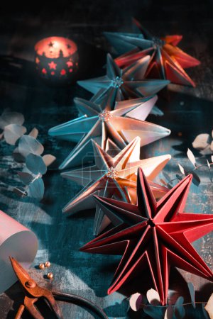 Photo for Handmade paper stars, self made Christmas decorations. Xmas ornaments on dark background. Origami craft hobby. Scissors, golden cord and glitter. Eucalyptus twigs and candles on dark wooden table. - Royalty Free Image