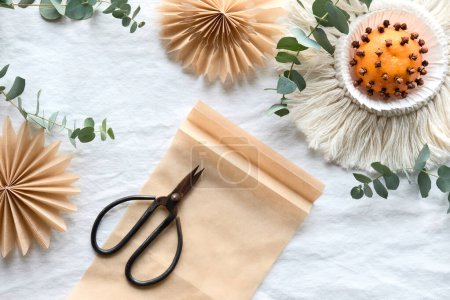 Photo for Fragrant pomander balls handmade from tangerines with cloves. Flat lay on off white textile tablecloth with aromatic wintertime green eucalyptus twigs. Old scissors on page of brown baking paper. - Royalty Free Image