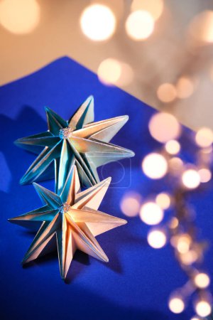Téléchargez les photos : Des étoiles en papier faites à la main, des décorations de Noël faites maison. Ornements de Noël sur fond de papier bleu foncé et jaune doré. Lumières brillantes, guirlande festive en bokeh. Origami bricolage passe-temps. - en image libre de droit